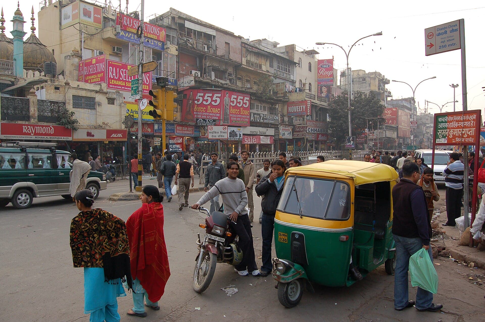 A green-and-yellow auto-rickshaw amid the crowds, saree-shop hoardings and tangled signboards of Chandni Chowk