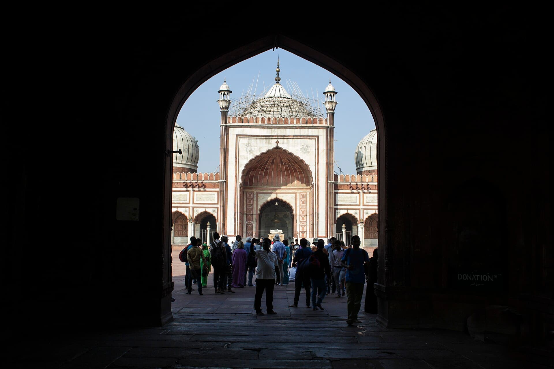 Jama Masjid's marble-domed prayer hall framed in silhouette through the great eastern gateway arch, worshippers in the courtyard