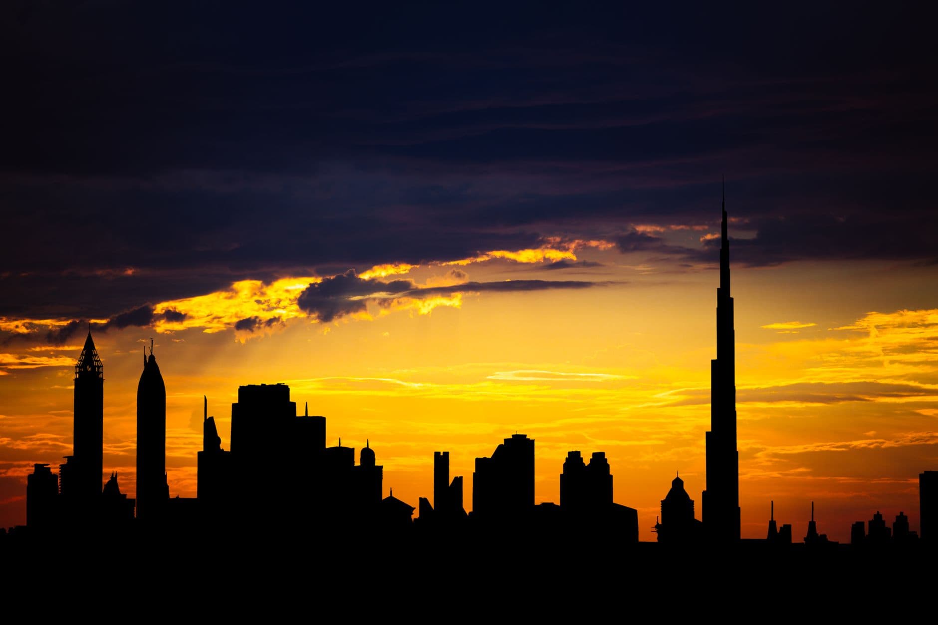 Dubai's skyline in silhouette at sunset, the Burj Khalifa's spire standing tallest against a golden-orange sky