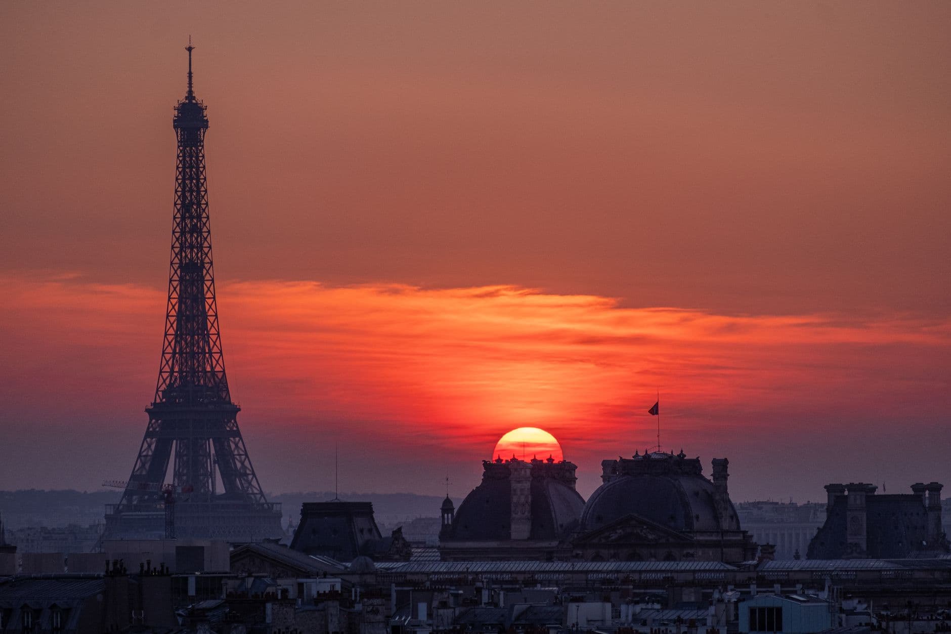 The Eiffel Tower silhouetted against a deep red sunset above the domed rooftops of Paris