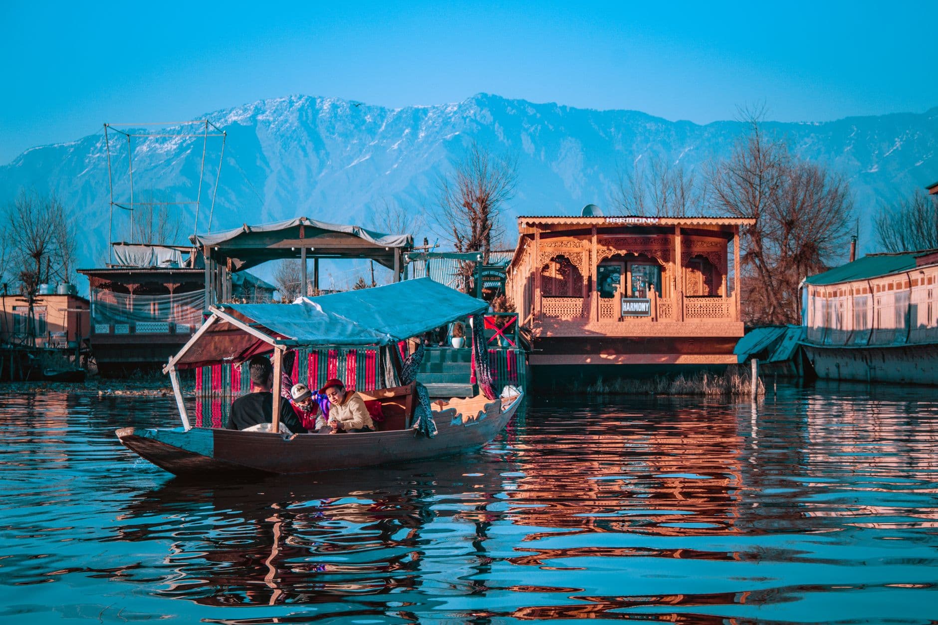 Tourists riding a canopied shikara past ornate wooden houseboats on Dal Lake at dusk, snow-dusted mountains behind