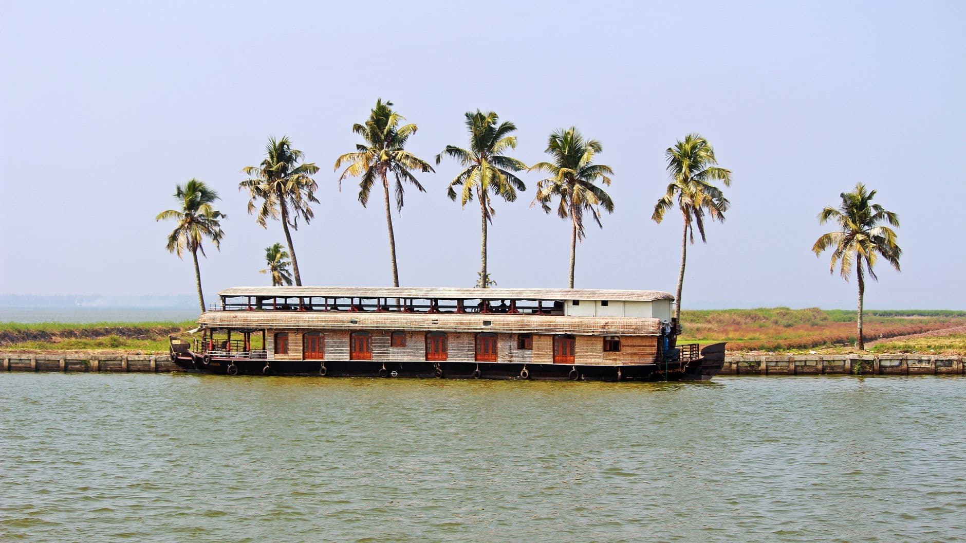 A two-deck kettuvallam houseboat moored along a palm-lined bank of the Kerala backwaters under a clear sky