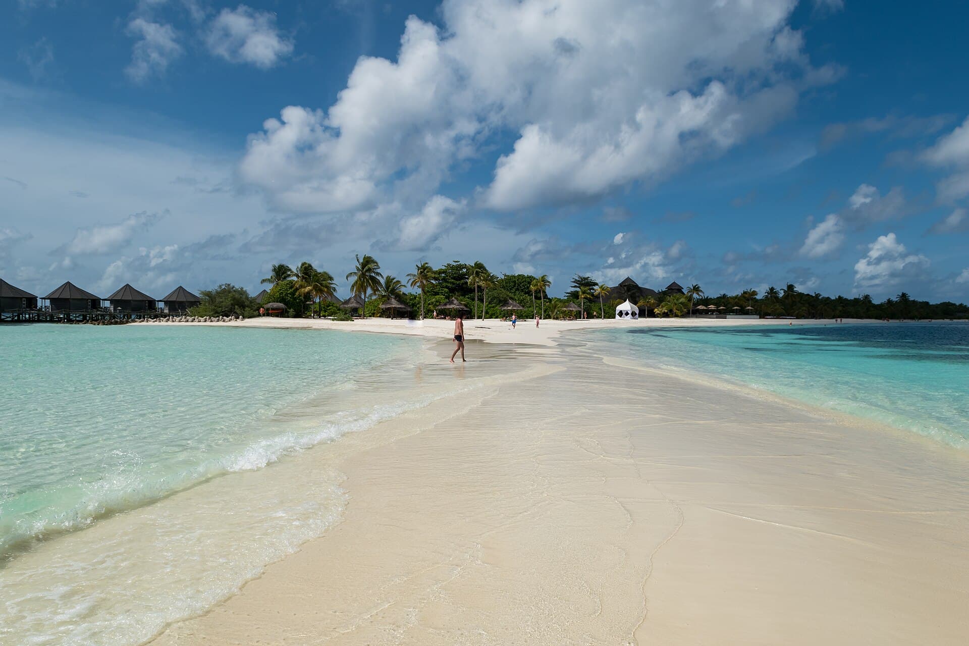 A person walking along a bare white Maldivian sandbank, overwater villas and palm trees visible behind