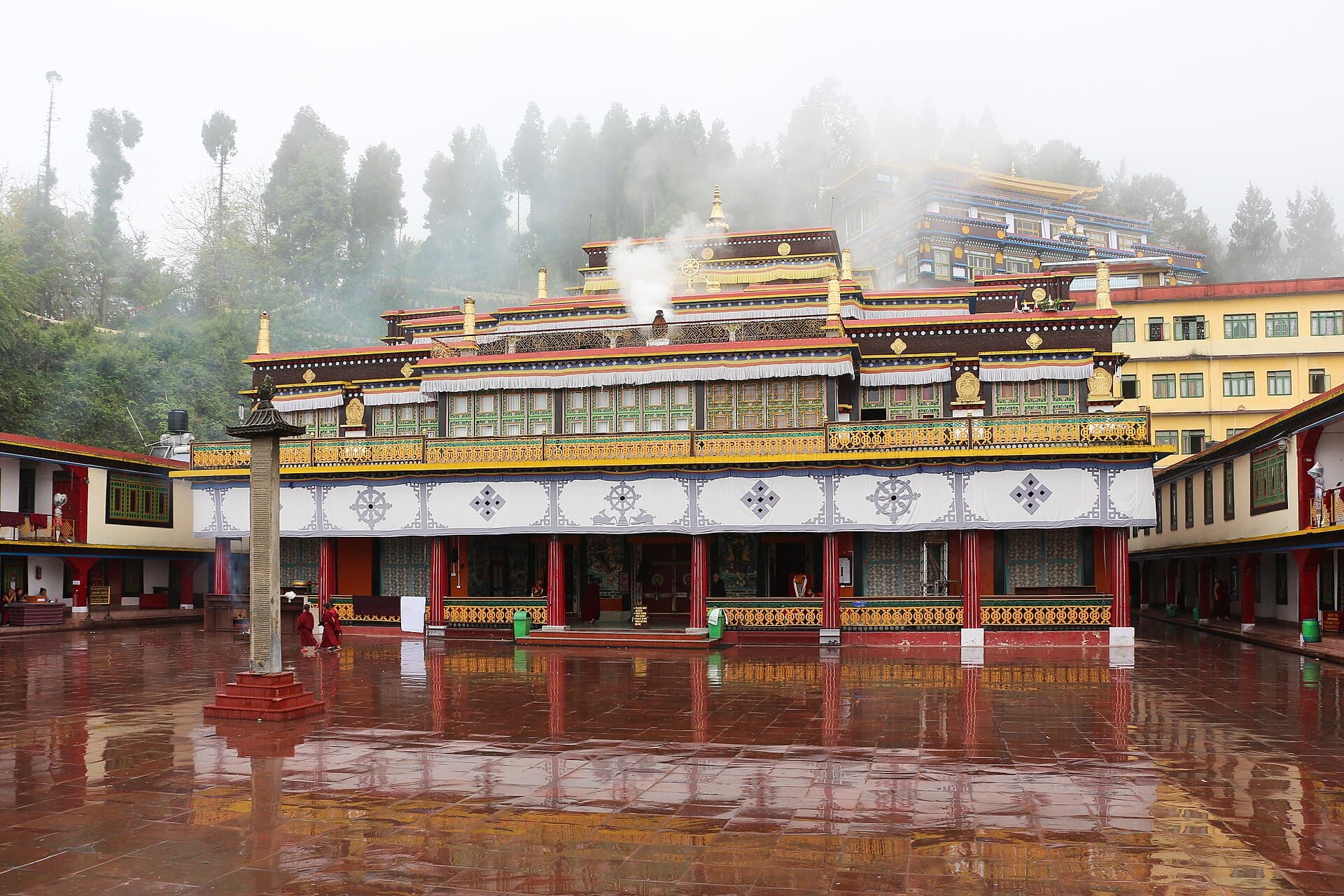 Rumtek Monastery's ornately painted main hall reflected in a rain-wet courtyard, monks in red robes crossing and mist in the trees