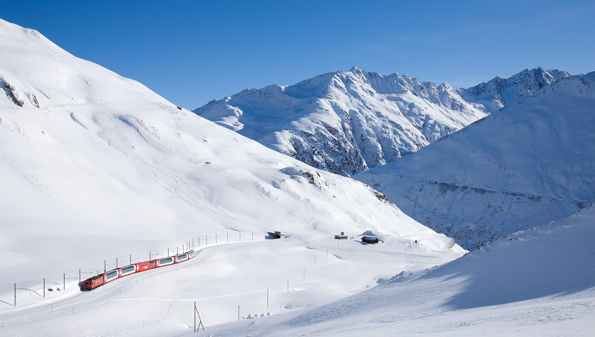 A red Glacier Express train winding through a snow-covered valley just below the Oberalp Pass summit