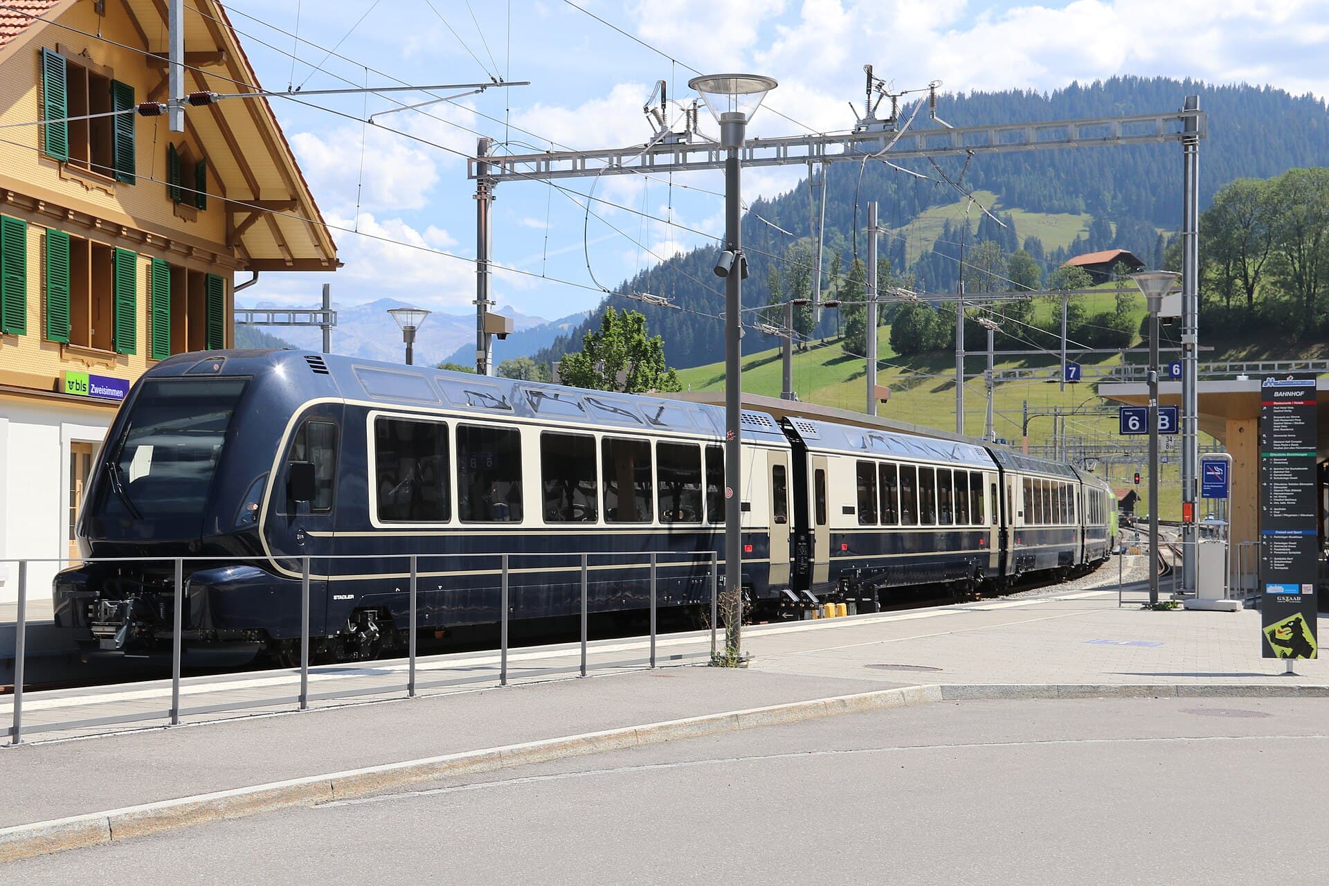 A dark blue GoldenPass panoramic train stopped at Zweisimmen station with green alpine meadows behind