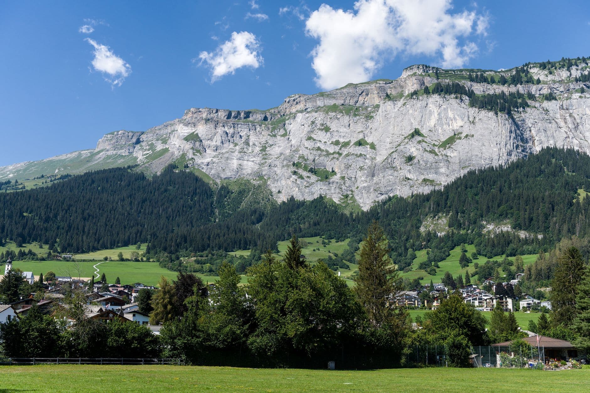 A Swiss Alpine village nestled beneath dramatic pale limestone cliffs under a blue sky with scattered clouds
