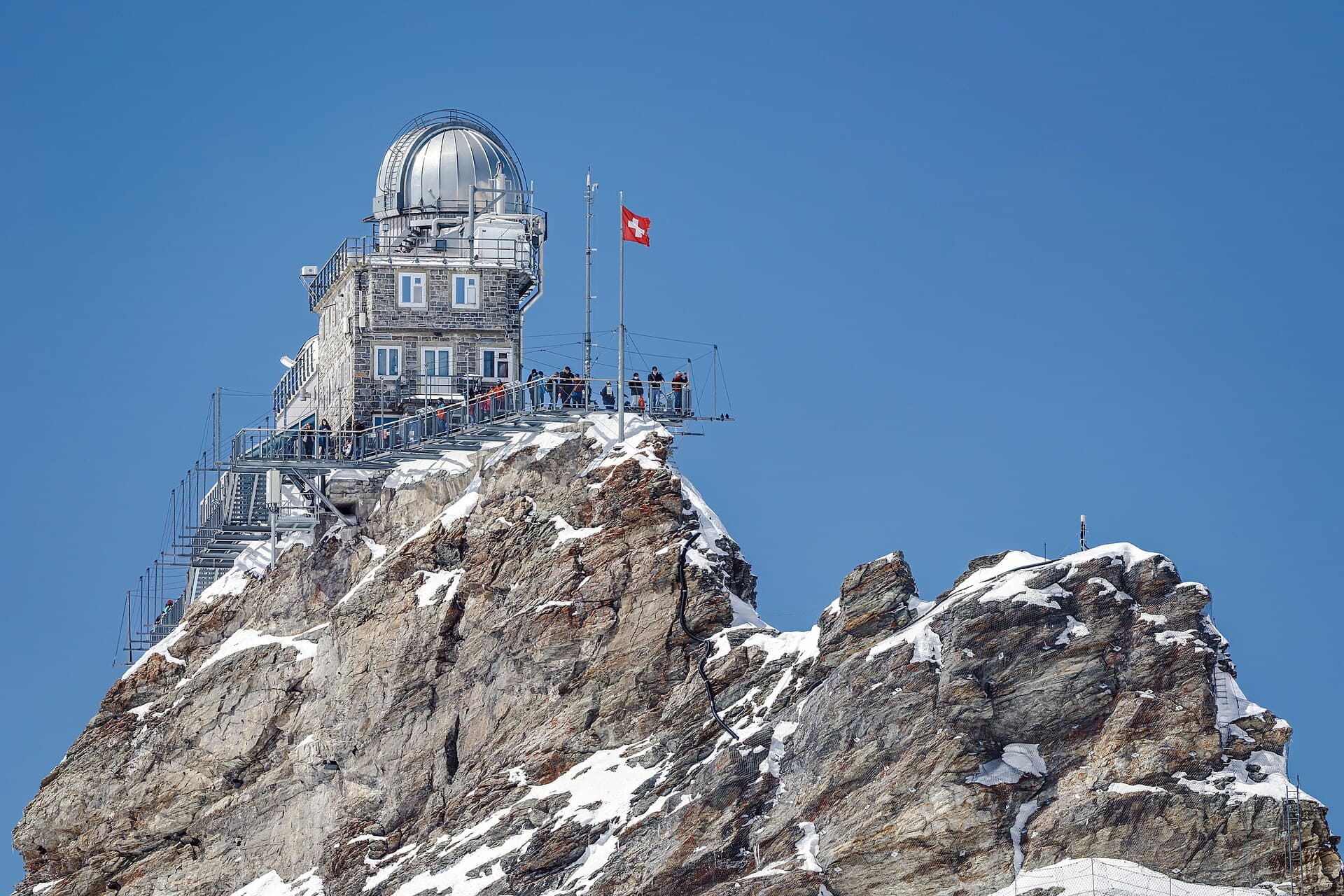 The stone Sphinx Observatory building perched on Jungfraujoch's rocky summit, a Swiss flag flying against a clear blue sky