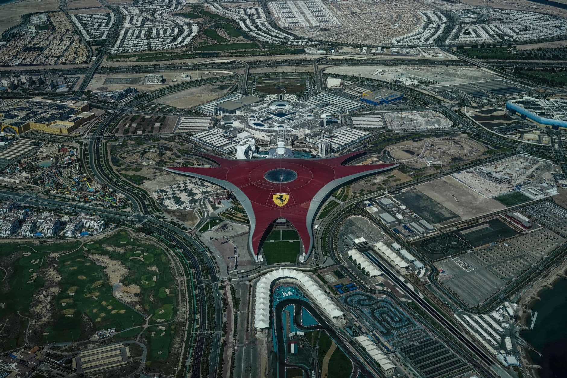Aerial view of Ferrari World's distinctive red spaceframe roof spread across Yas Island, race track curling beneath it