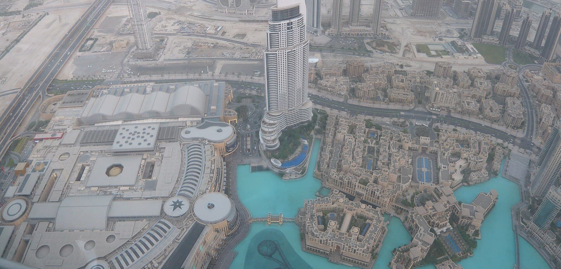 Aerial view of Dubai Mall's curved rooftop and the turquoise Burj Khalifa Lake fountain beside it