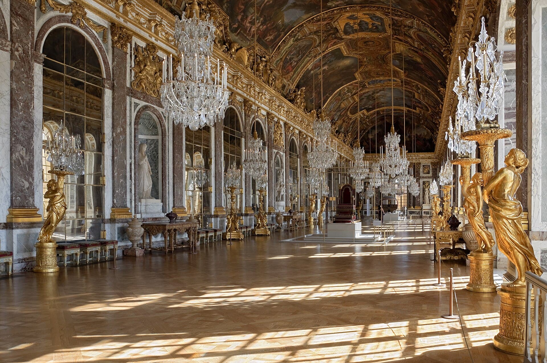 The Hall of Mirrors at Versailles, its crystal chandeliers and gilded statues lit under a frescoed vaulted ceiling
