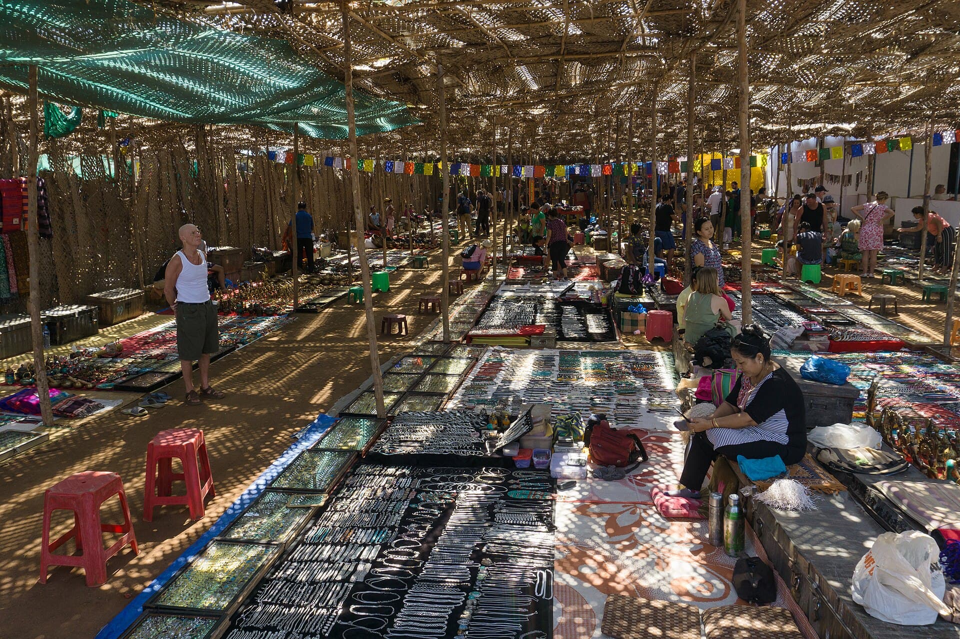 Rows of jewellery and handicraft stalls under Anjuna Flea Market's woven-palm canopy, dappled sunlight falling on browsing shoppers