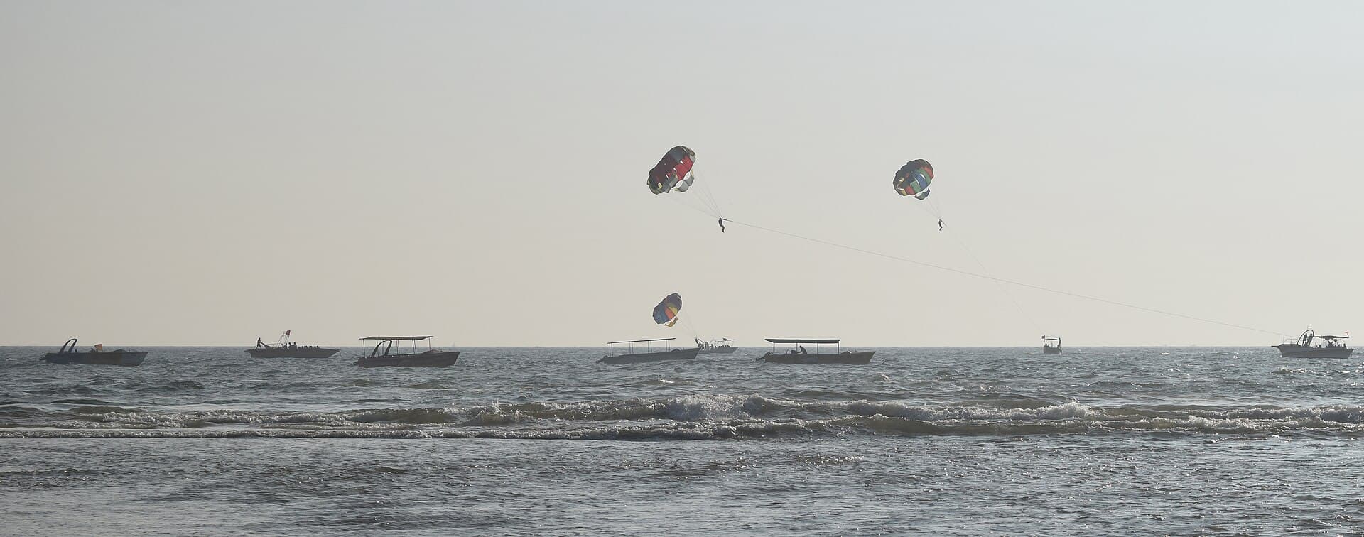 Three parasailers being towed above water-sports boats in the surf off Baga Beach in the late-afternoon haze