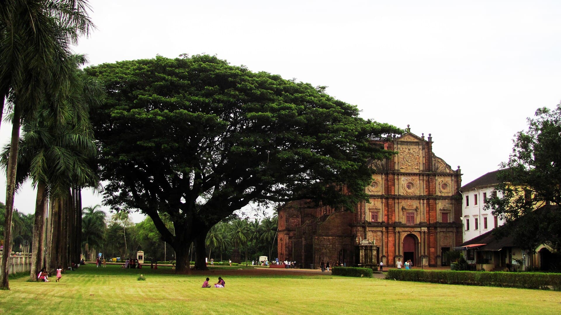 The red laterite facade of the Basilica of Bom Jesus in Old Goa rising behind a huge rain tree, with visitors resting on the lawn in front