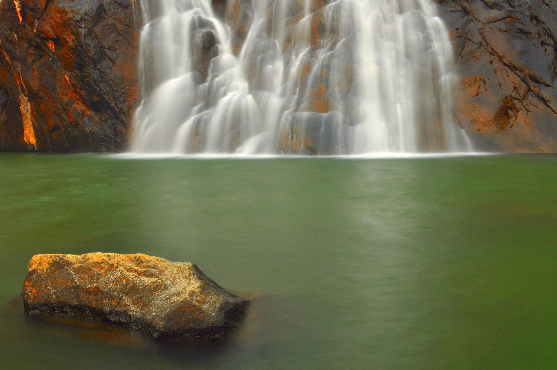 White cascades of Dudhsagar Falls pouring down dark rock into a still green plunge pool, a sunlit boulder in the foreground