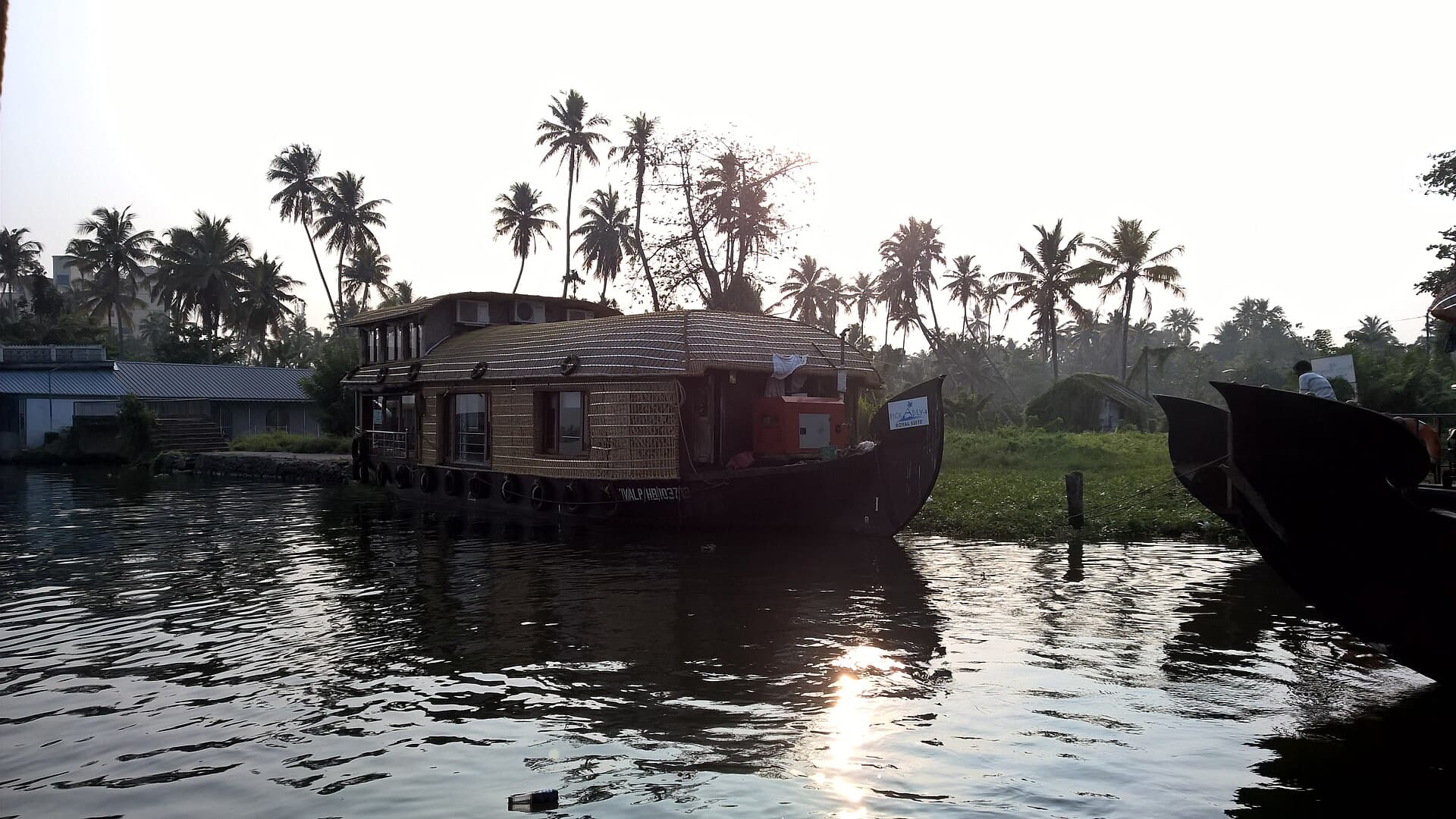 A thatched houseboat on the Alleppey backwaters at sunset, coconut palms silhouetted behind and sunlight glinting off the water