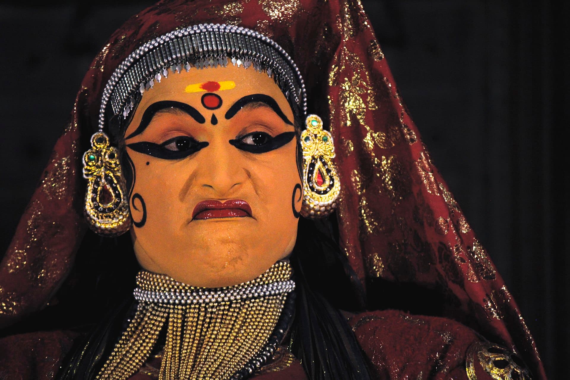 Close-up of a Kathakali performer in a female role — painted face, bold black eye make-up, silver headdress and heavy jewellery