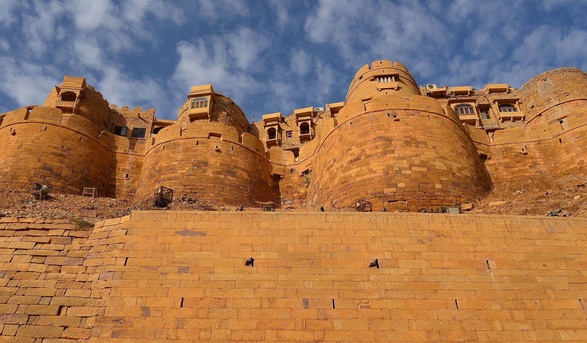 The round golden-sandstone bastions of Jaisalmer Fort rising from their sloped stone base against a blue desert sky