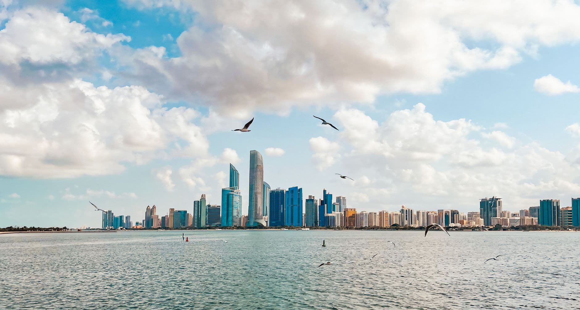Abu Dhabi's skyline of glass towers seen across the water from the Corniche, seagulls flying overhead under a cloudy sky